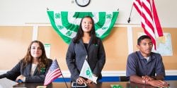 Three youth behind a desk with banners and national flag; Alabama 4-H