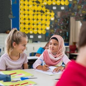Young arab girl with hijab doing exercise with her bestfriend at international school. Asian muslim school girl sitting near her classmate during lesson. Multiethnic elementary students in classroom.