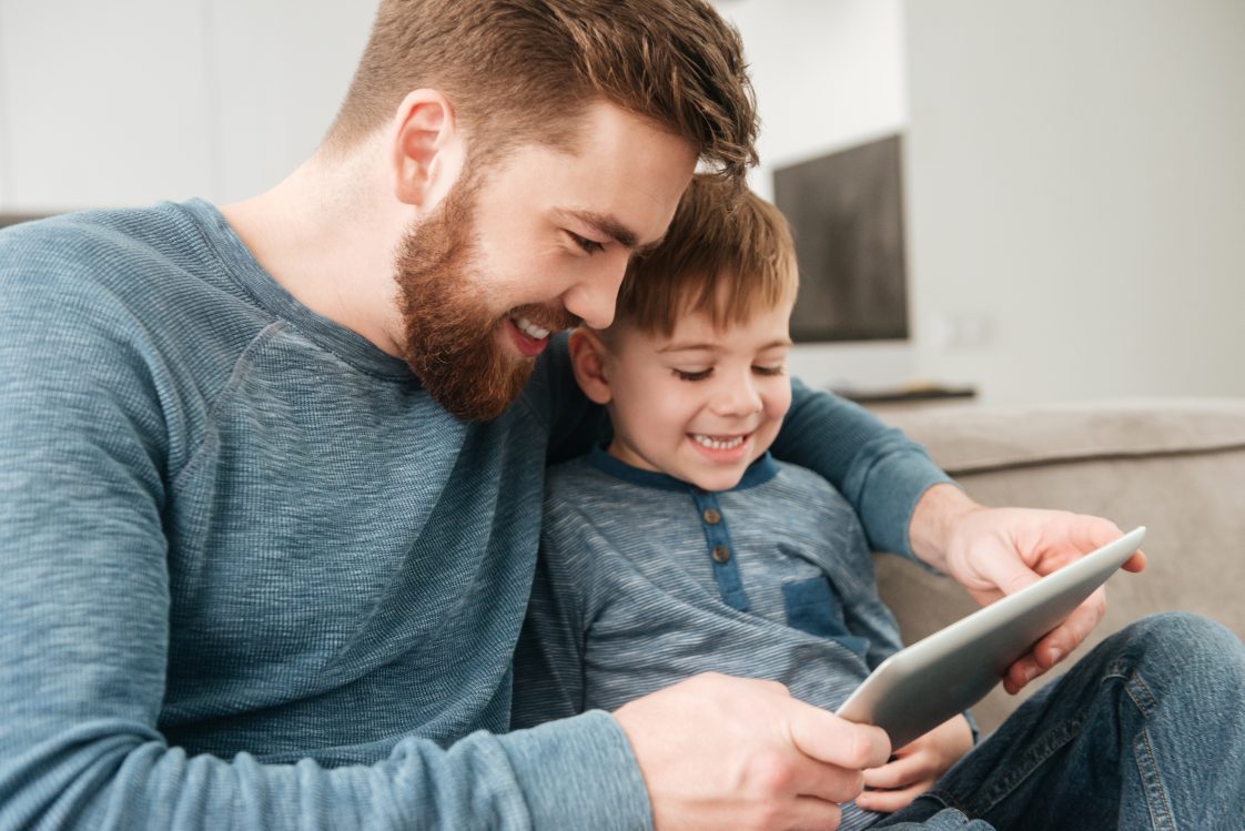 A father and son sitting on a couch looking at a tablet.