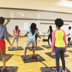 The exergame screen is projected on the gym wall as a group of school-age children use there exergame controller matts to play.