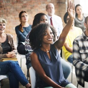 Group of people sit a room. One woman holds her hand up to volunteer