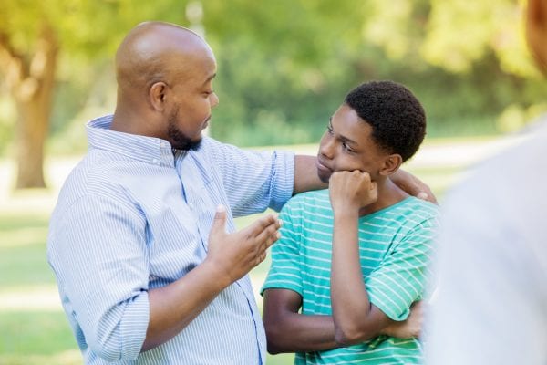 An African American father stands outdoors with his teenage son and places a hand over his shoulder as he gestures and shares some bad news. An unrecognizable person looks on as the son listens to his father with a gesture of disappointment.