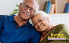 An Indian American older adult couple sits on their sofa, smiling.