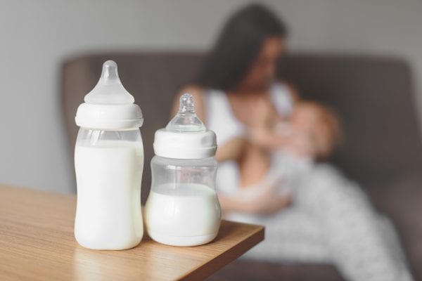 Bottles with breast milk on the background of mother holding in her hands and breastfeeding baby. Maternity and baby care.