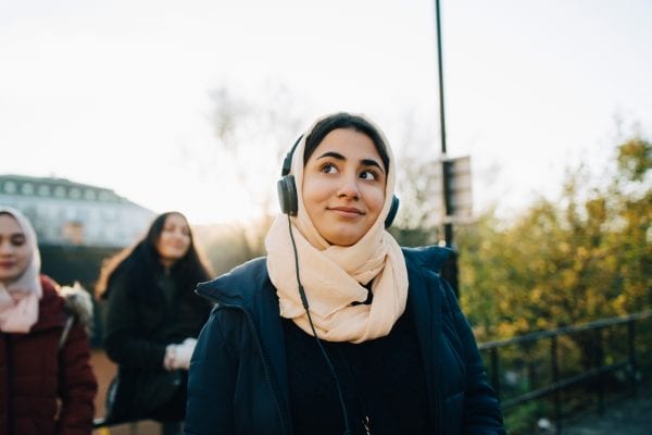 Smiling teenage girl listening to headphones with friends against sky
