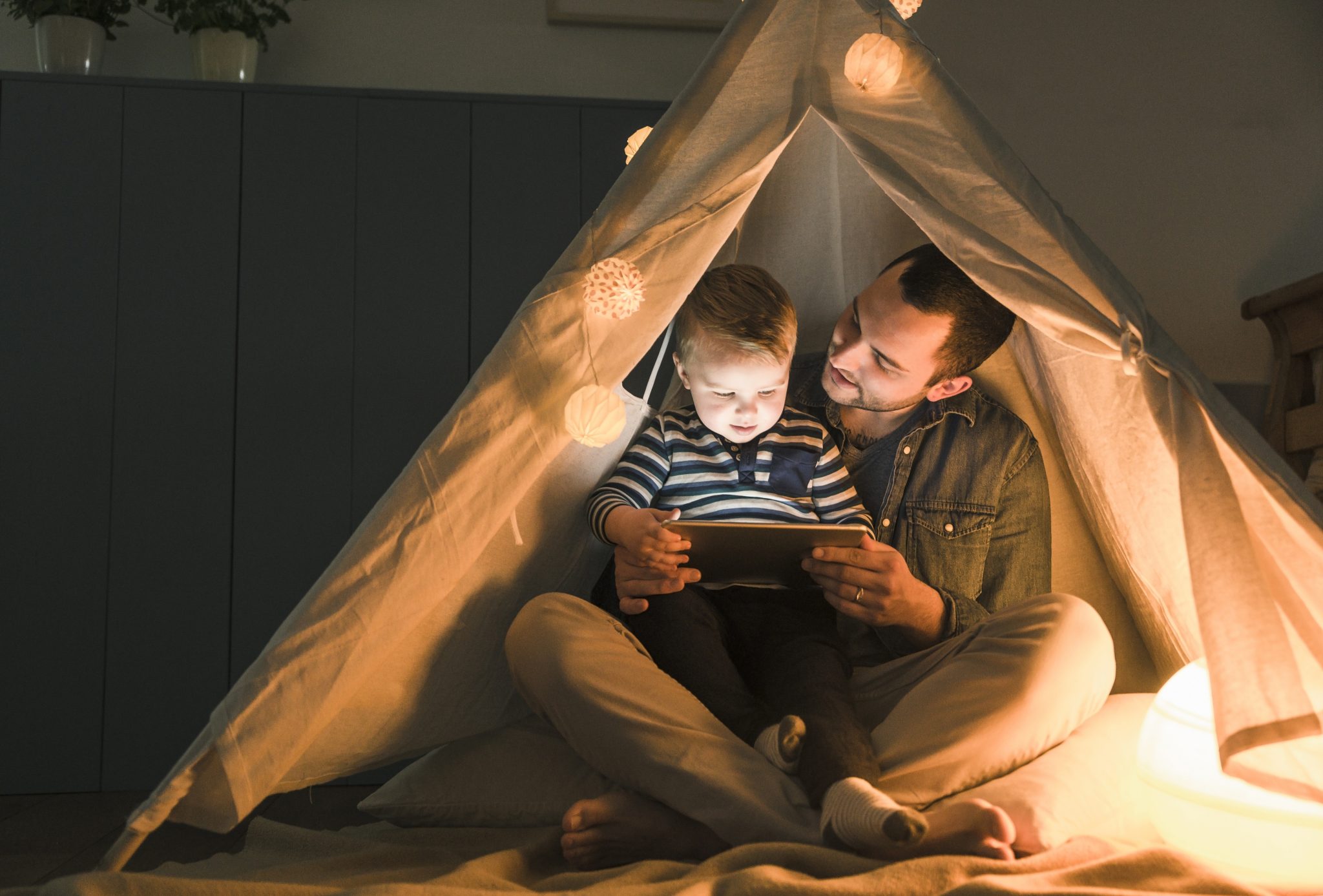 Father and son sharing a tablet in a dark tent at home