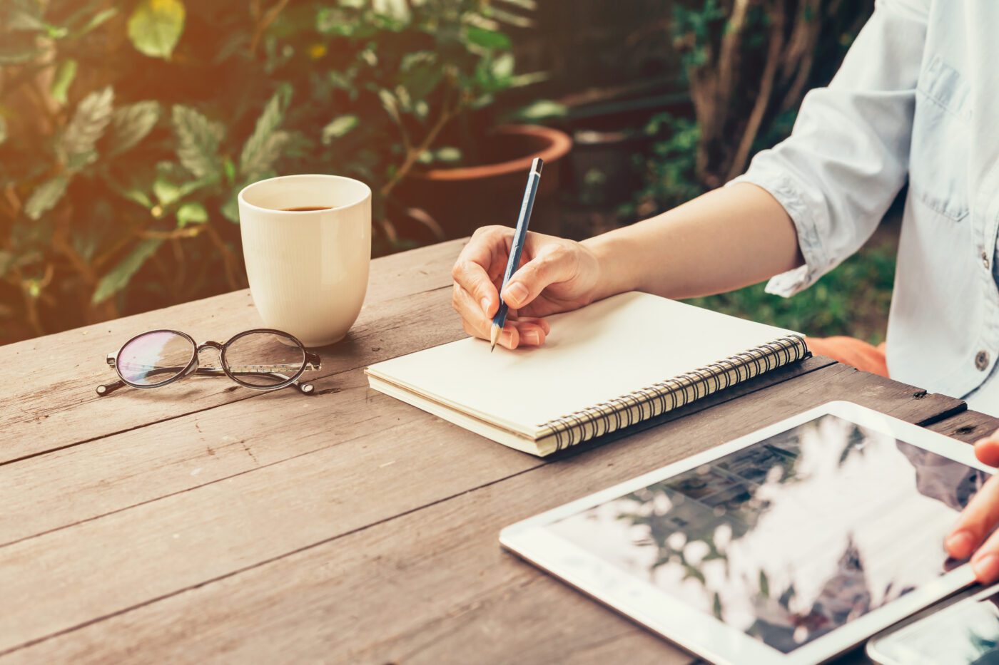 a person journaling outside. image shows their hand holding a pencil over a journal with a cup of coffee and glasses next to it