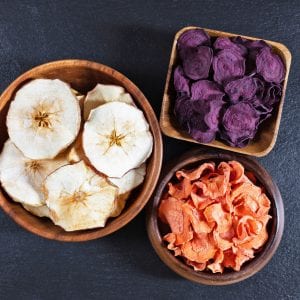 Dried carrot, beetroot and apple chips in a wooden bowl on a black stone background.