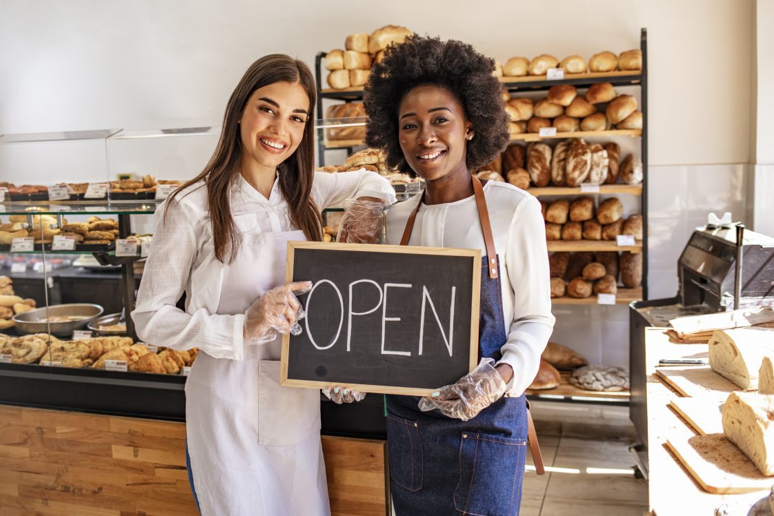 Happy businesswomen standing at bakery shop with open signboard. Close up of women holding sign now we are open support local business. Happy business owner welcoming customers at a bakery shop