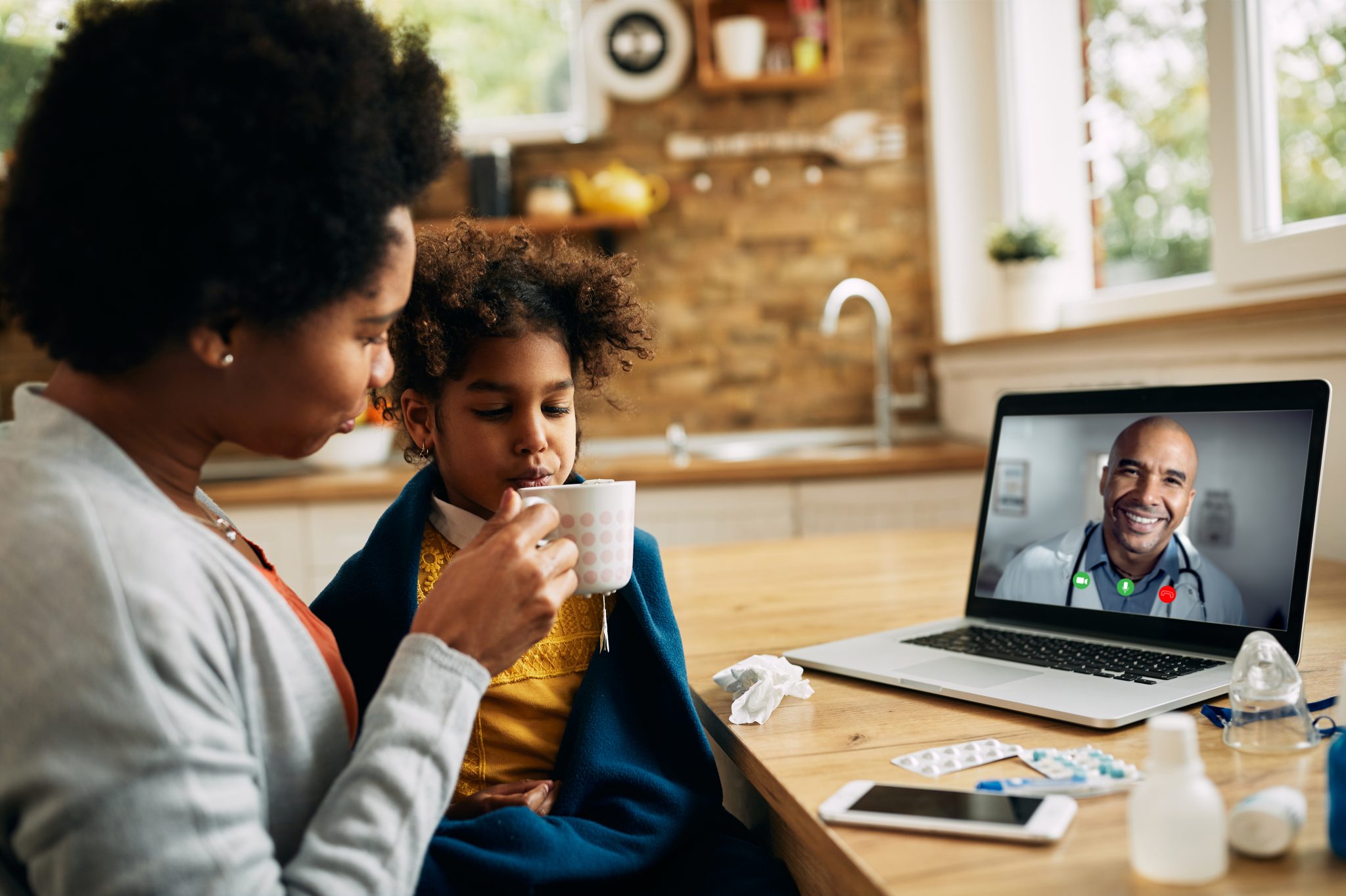 Caring African American mother giving tea to her ill daughter during video call with a pediatrician at home.