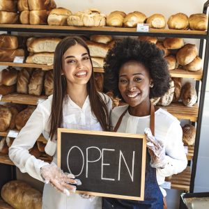 Stop by for homemade delicious bread! Loving young women embracing holding a sign together on the opening day of their small business - couple owners of the bakery smiling to the camera