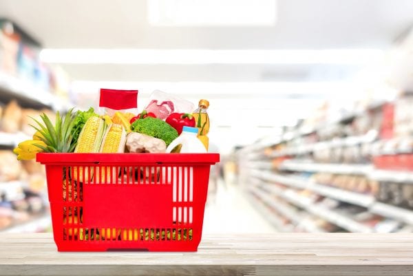 Food and groceries in red shopping basket on wood table with blurred suppermarket aisle in background
