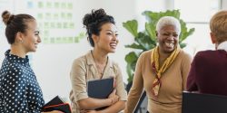 Colleagues standing in a small group discussing something while laughing. Two of the women are holding notebooks.