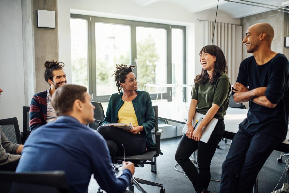 Colleagues looking at cheerful businesswoman holding laptop in meeting at creative office