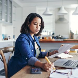 Shot of a young woman using a laptop and going through paperwork while working from home