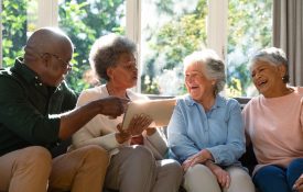 Three happy diverse senior woman and african american male friend sitting on sofa and using tablet. retirement lifestyle relaxing at home with technology.