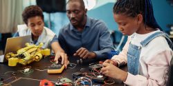 Side view portrait of young black girl building robots with male teacher in background during engineering class at school