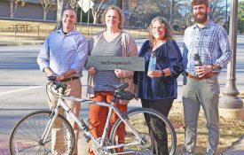 ALProHealth team members Jeffrey LaMondia, Ruth Brock, Sondra Parmer, and Mitch Carter stand on sidewalk holding sign and bicycle.