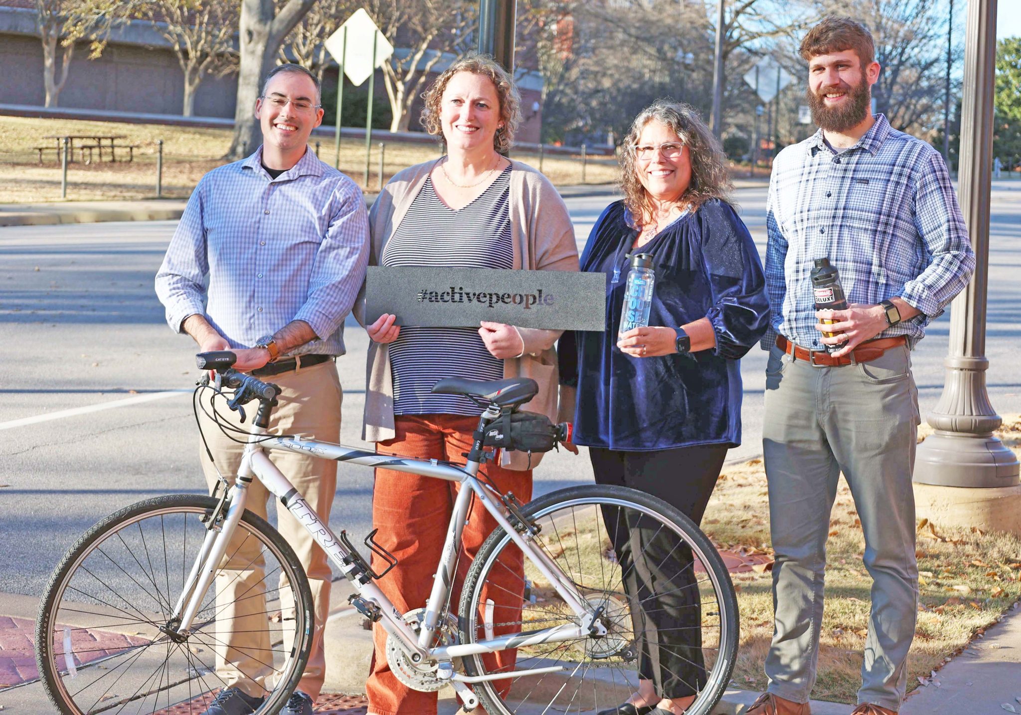 ALProHealth team members Jeffrey LaMondia, Ruth Brock, Sondra Parmer, and Mitch Carter stand on sidewalk holding sign and bicycle.