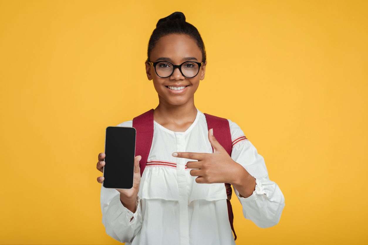 Satisfied smart adolescent black lady pupil in glasses with backpack shows finger at phone with blank screen, isolated on yellow background. App for study, online device, website for remote lesson