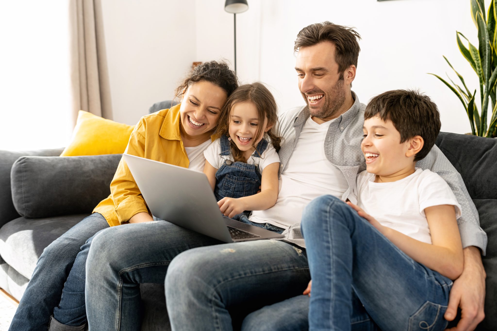 Cheerful happy family of four using laptop sitting on the sofa in cozy apartment, parents and two kids watching movie on computer, comedy, spends time on the weekend together at home