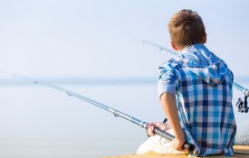 A boy in a blue plaid shirt sitting on a dock with a fishing pole.