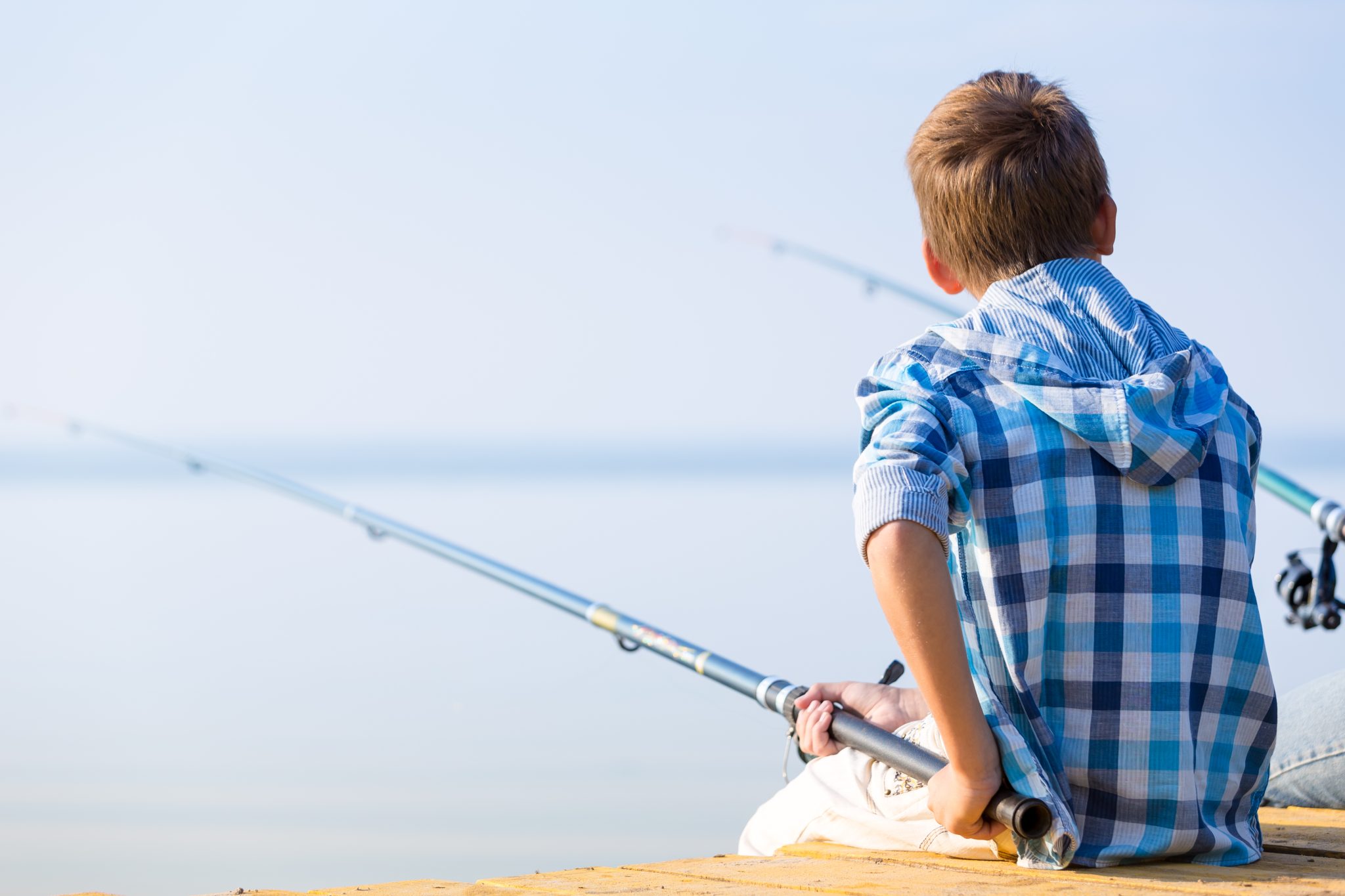 A boy in a blue plaid shirt sitting on a dock with a fishing pole.