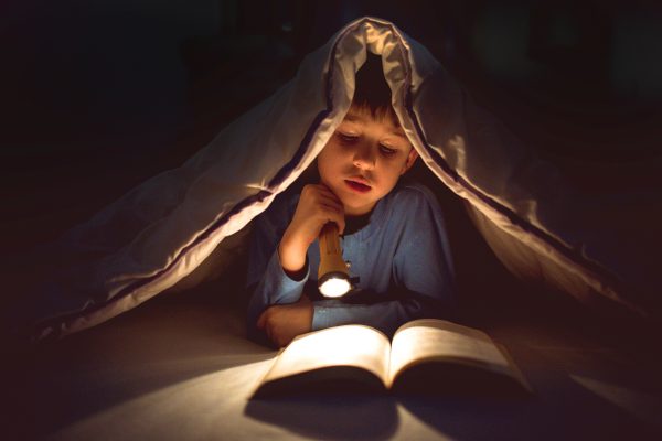 Little boy reading a book under the covers with flashlight