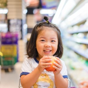 Funny little child smile and holding apple.Asian kid Shopping in supermarket.Little asian toddler girl sitting in the trolley during family shopping in hypermarket.