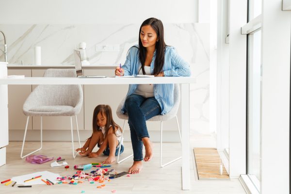 Young Pretty Asian Woman Working With Laptop And Notepad While Her Daughter Plays on the Floor