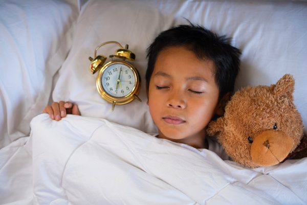 asian boy sleeping on bed white pillow and sheet with alarm clock and teddy bear.boy sleeping in morning