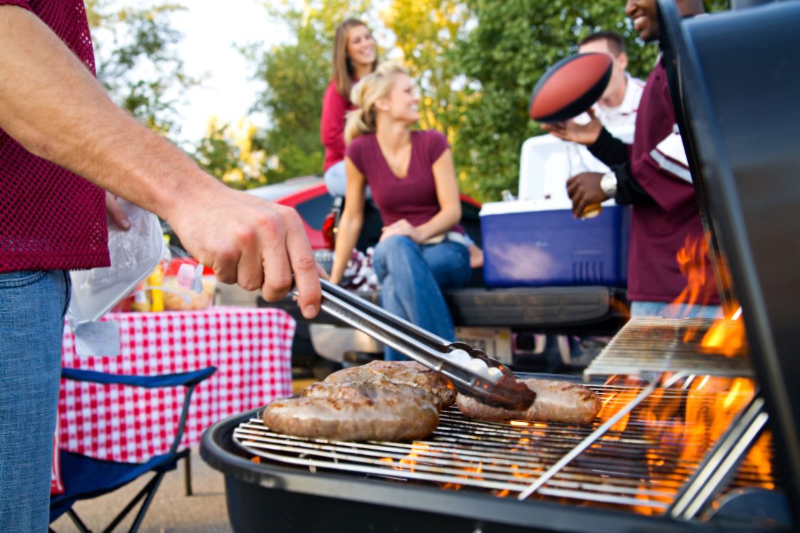 People in maroon shirts using a grill at a tailgate.