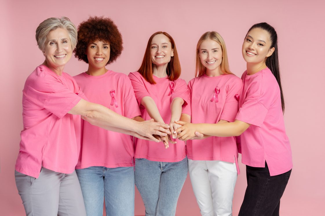 Group of smiling multiracial women with pink ribbon holding hands together. Breast cancer awareness