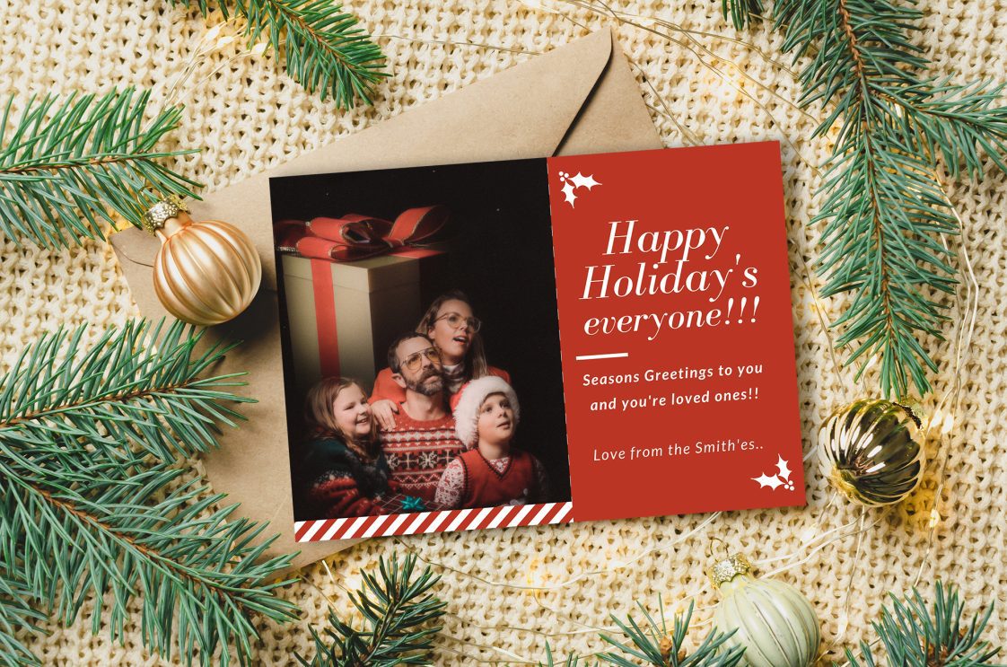 A holiday card with poor grammar and a bad family photo sitting on top of an envelope, surrounded by pine needles, ornaments, and lights.