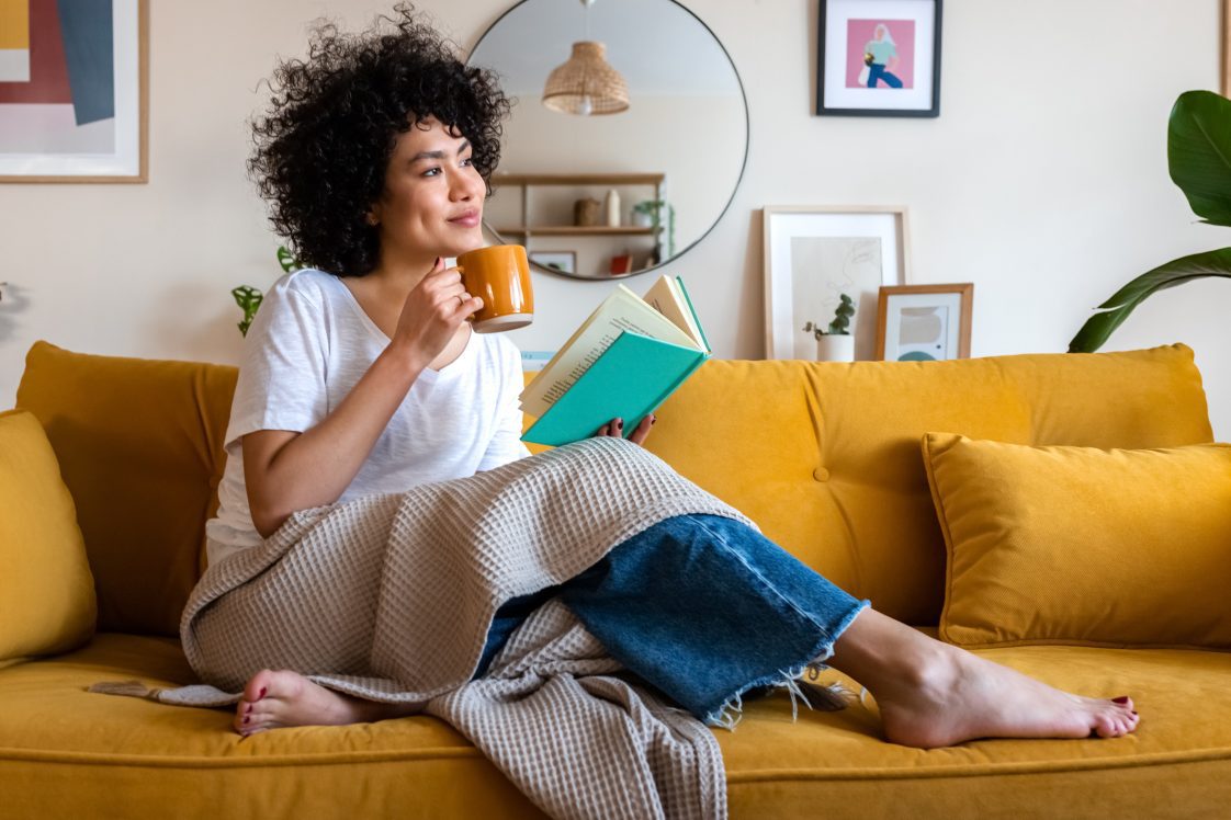 woman reading a book at home, drinking coffee sitting on the couch