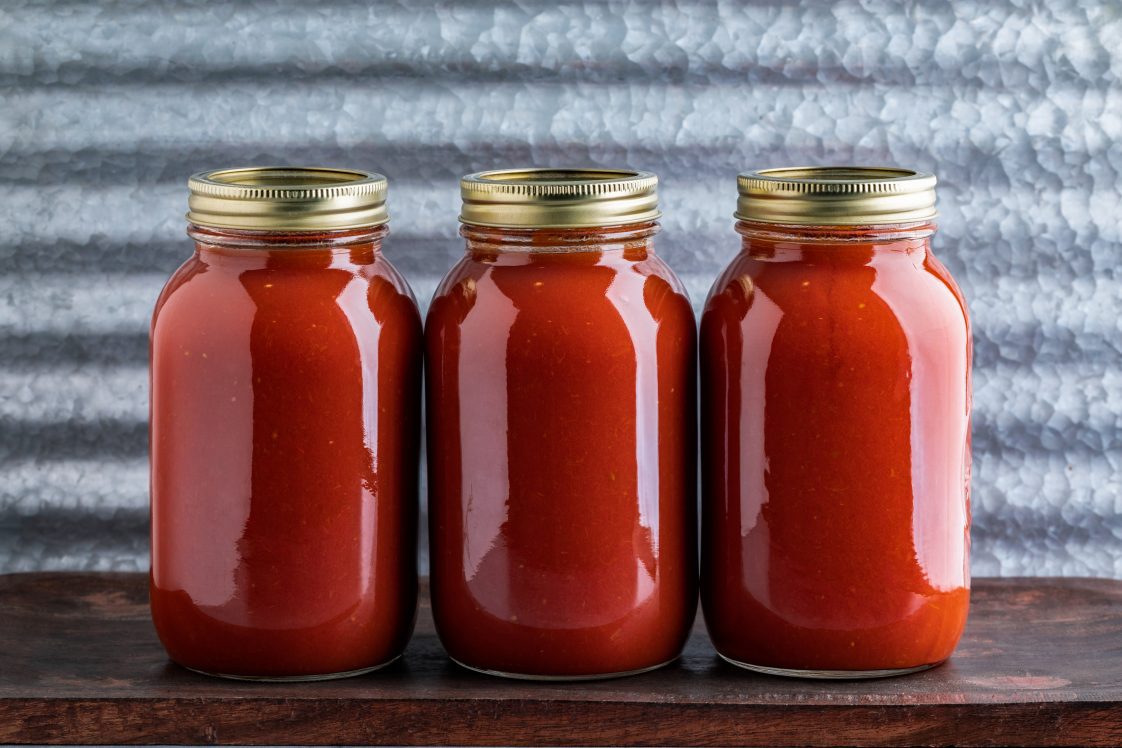 Three jars of homemade water bath canned crushed tomato sauce made from homegrown tomatoes on a wooden table and industrial background