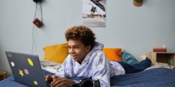 Smiling teenage boy relaxing on bed in front of laptop at leisure and playing games or communicating with friends in video chat
