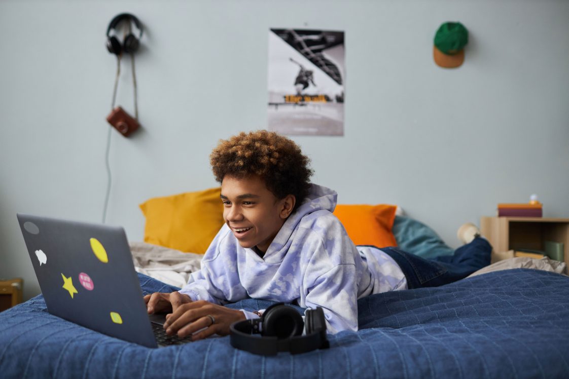 Smiling teenage boy relaxing on bed in front of laptop at leisure and playing games or communicating with friends in video chat