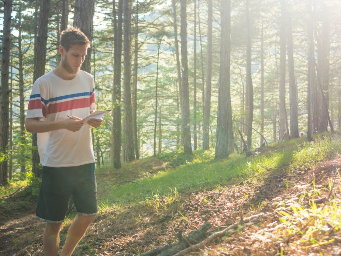 A younger man hold paper while walking through a forest.