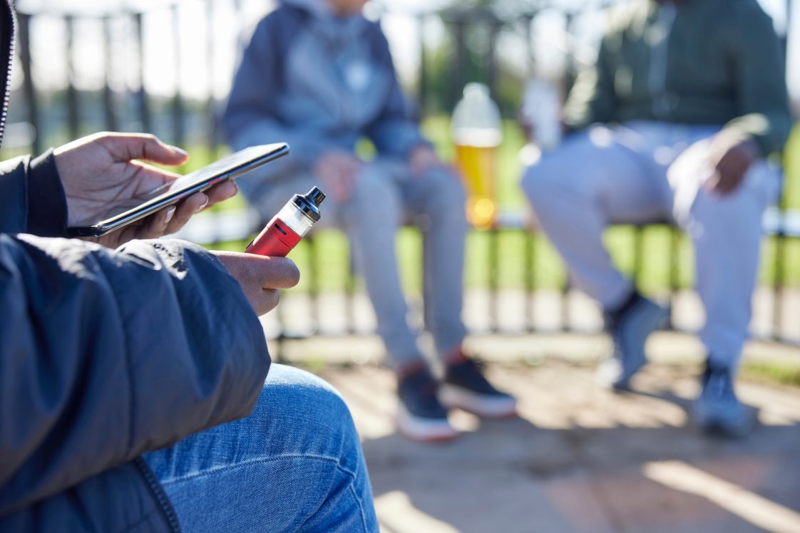 Close Up Of Teenagers With Mobile Phone Vaping and In Park