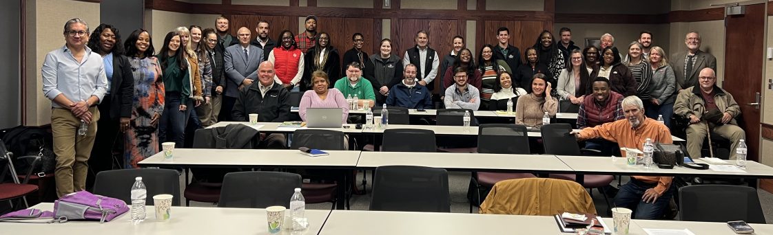 Photo of large group of people standing and sitting in a conference room setting.
