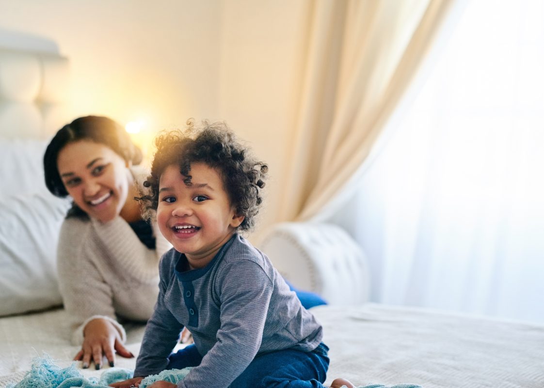 A young Black mother and her toddler playing on the bed.