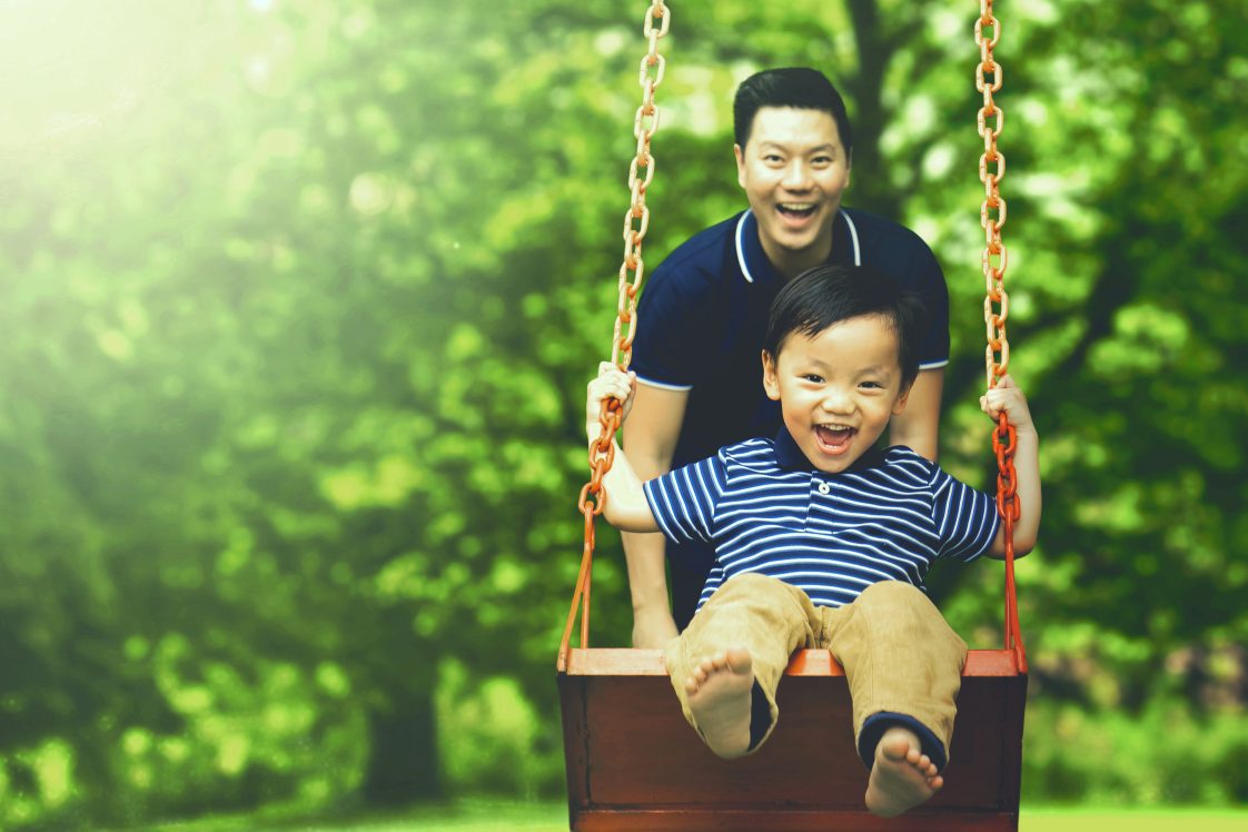 Image of young father pushing his son on the swing while having fun in the park.