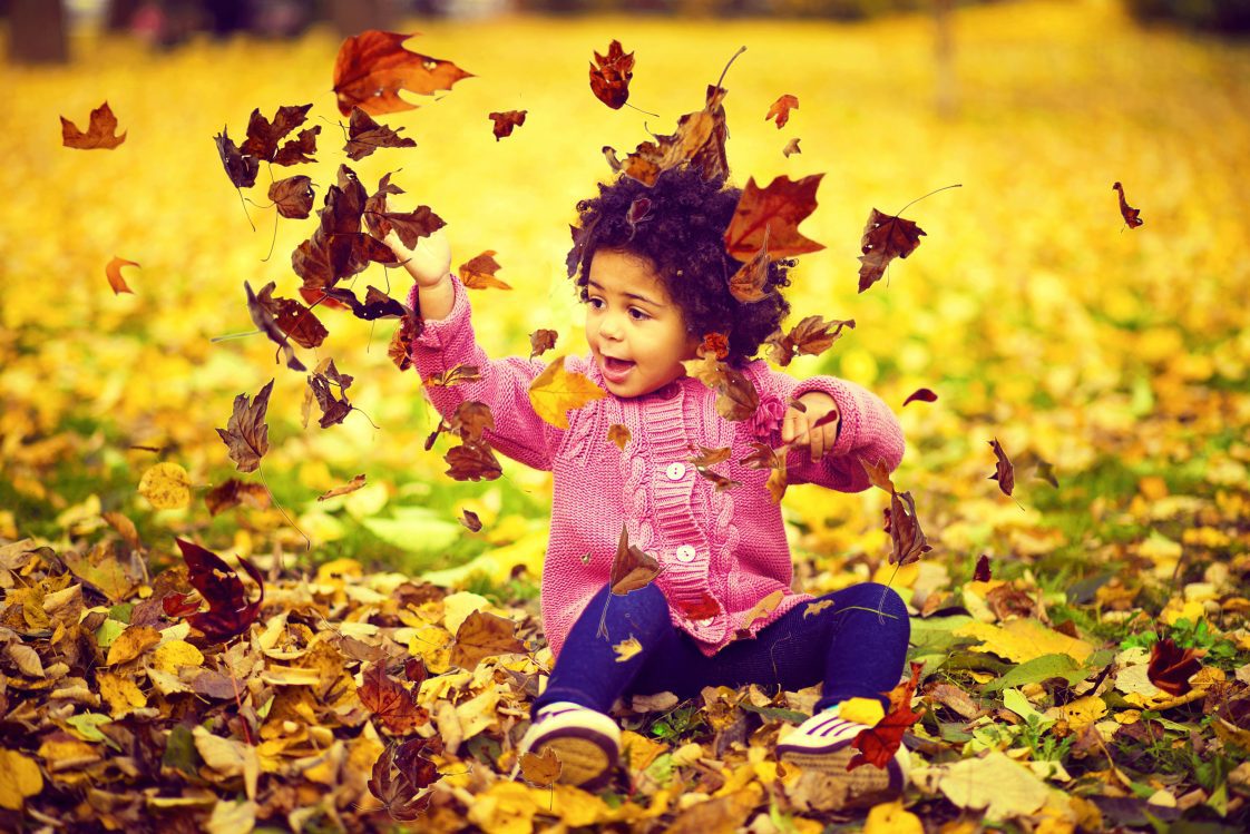 A young African American girl playing in the leaves on a fall day.