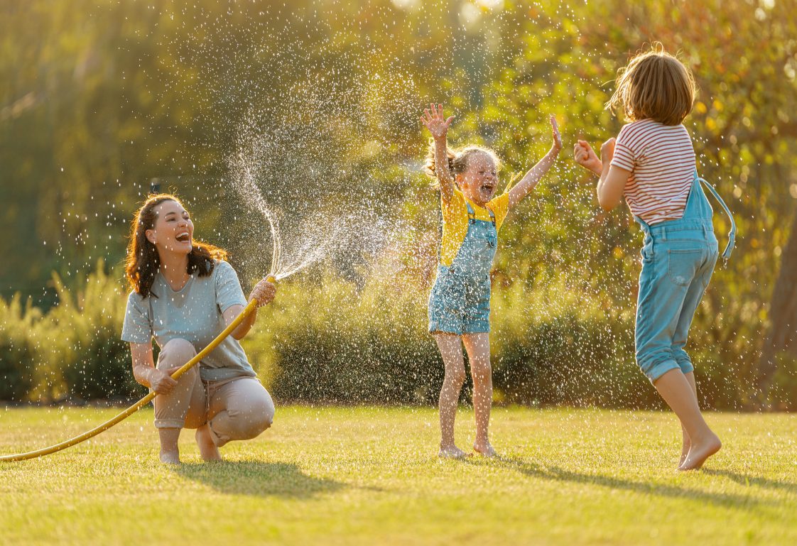 A mother and two daughters playing in the yard, with the mother spraying the girls with a water hose.