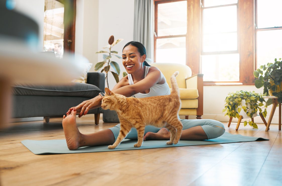 A young Black woman sitting on a yoga mat in her living room and petting an orange cat.