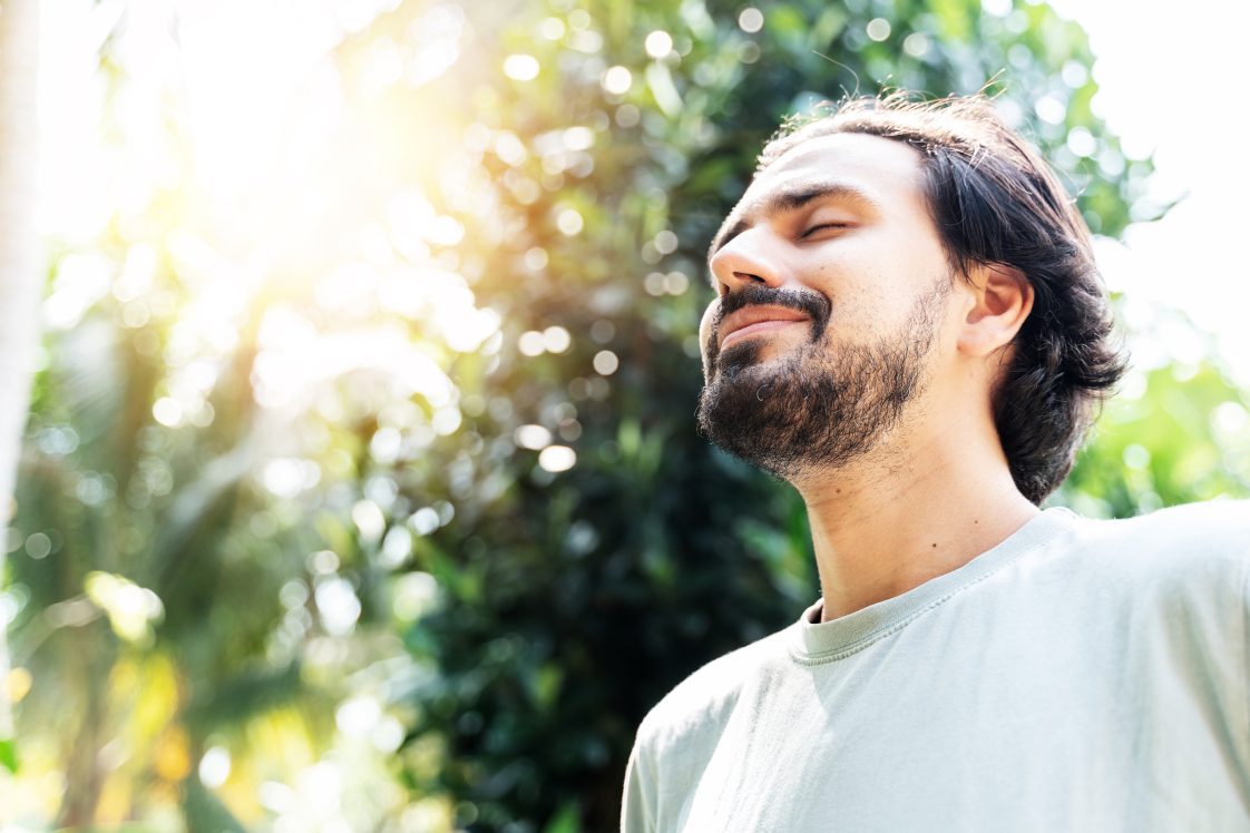 A bearded man is meditating outdoor in the park with face raised up to sky and eyes closed on sunny summer day.