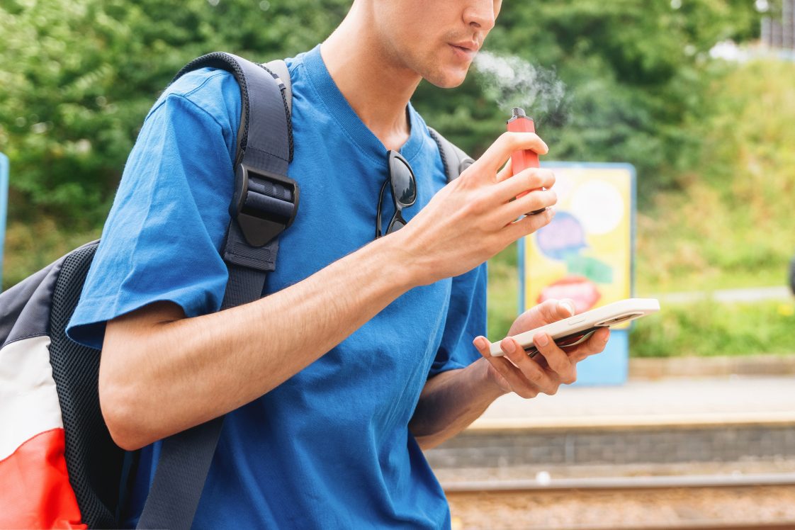 young man wearing a backpack and looking at his phone while using a vape device