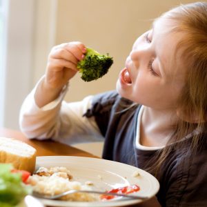 A 4 year old girl sitting at the table enjoying a healthy lunch. She is eating broccoli with her hands, her mouth is wide open. Soft window lighting.