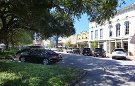 Cars parked in front of businesses along a shady street of a downtown square.